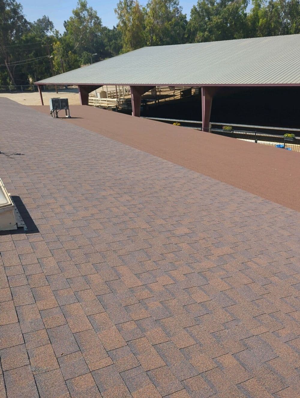 Newly installed asphalt shingles on a large flat roof with trees and structure in background.