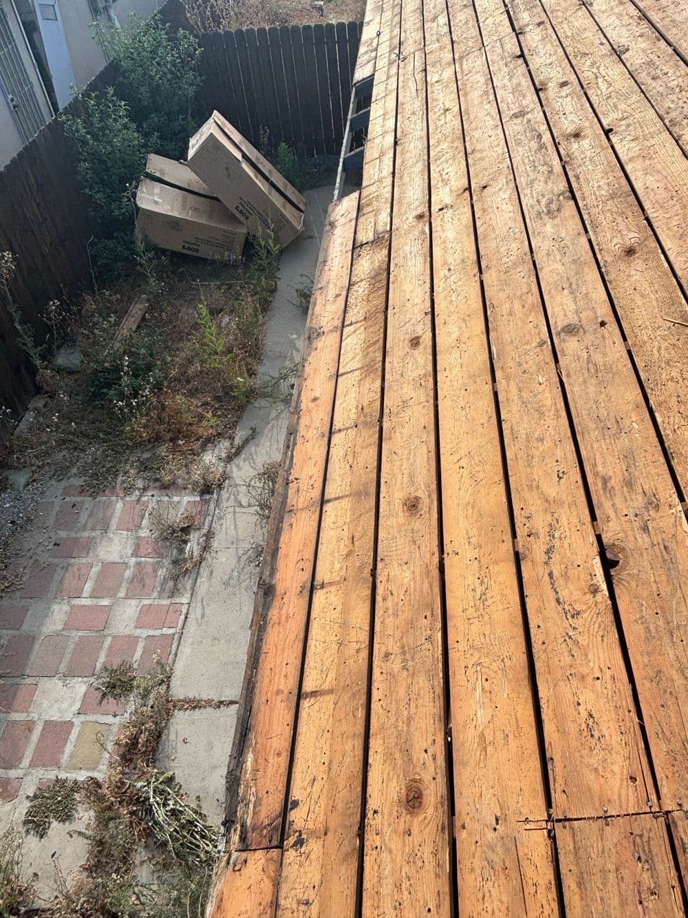 Wooden deck viewed from above, with scattered boxes and overgrown plants in the background.