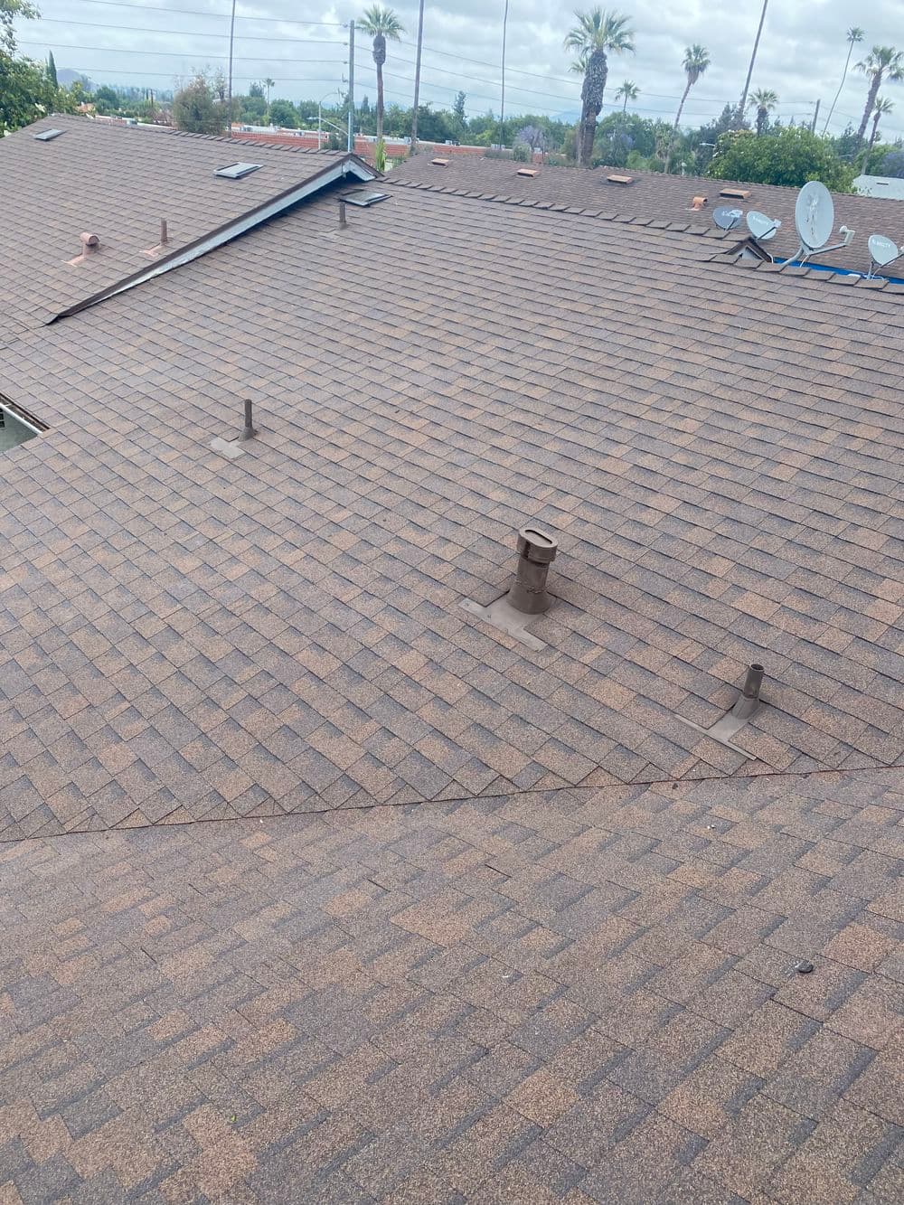 Roof with asphalt shingles, multiple vents, and satellite dishes, under a cloudy sky.