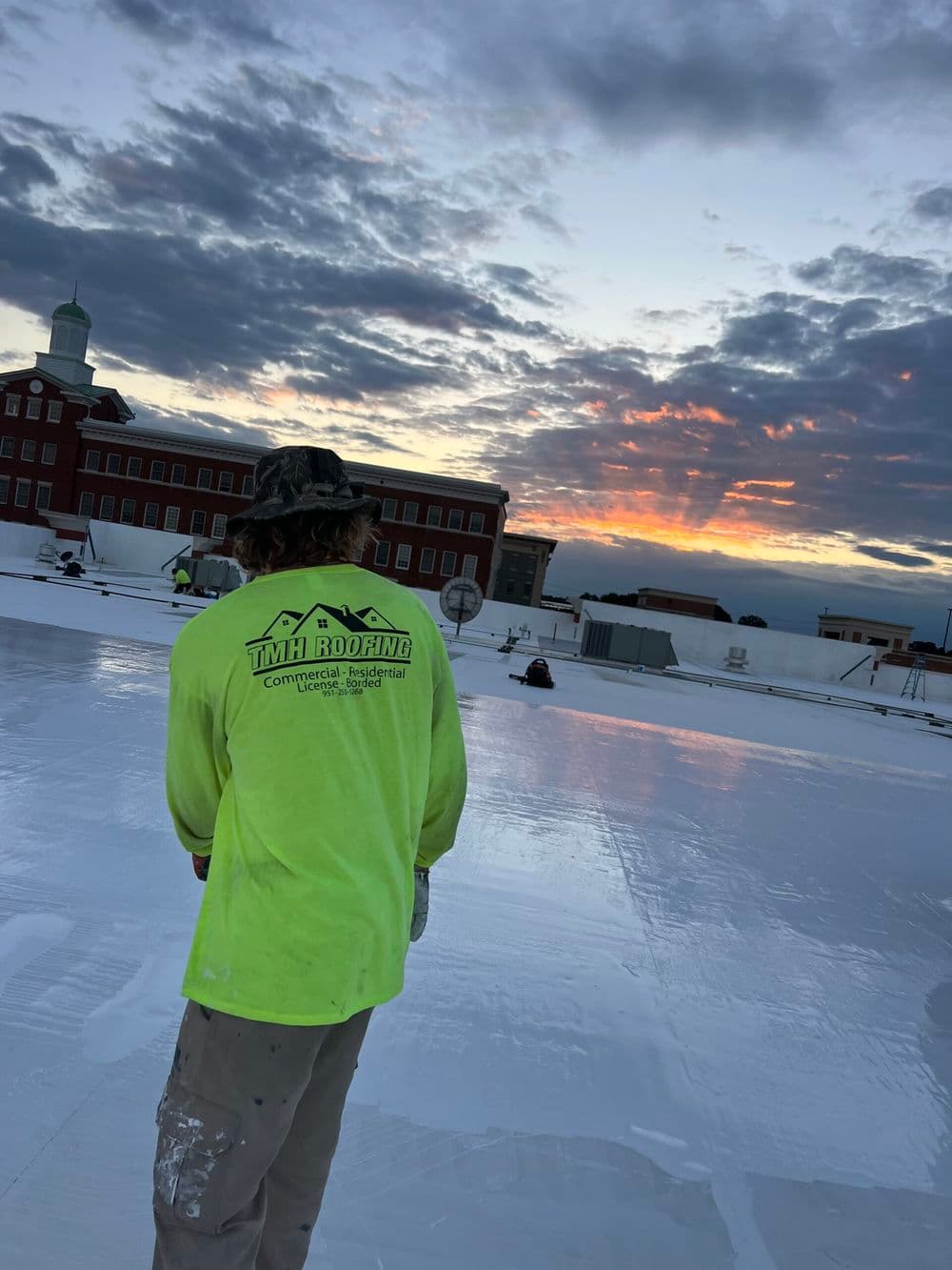 Roofer in a neon shirt works on a white rooftop at sunset with clouds and a building in background.